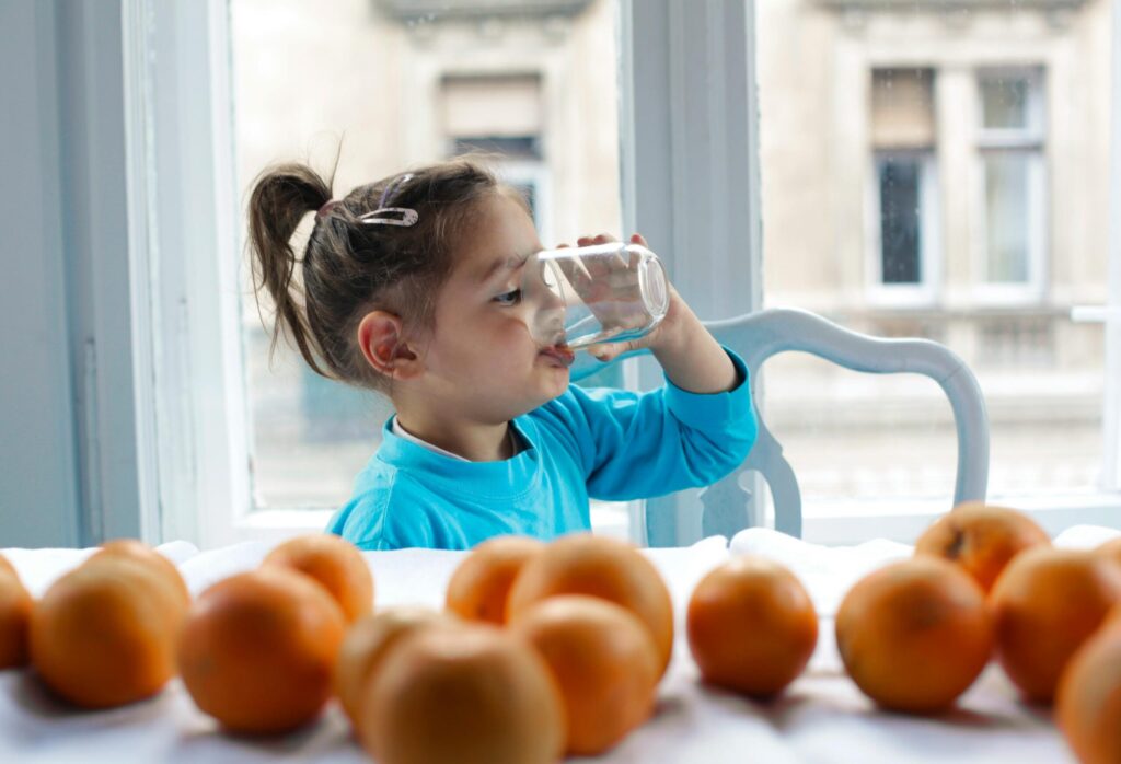 Child in blue sweater drinks water by a window surrounded by fresh oranges.